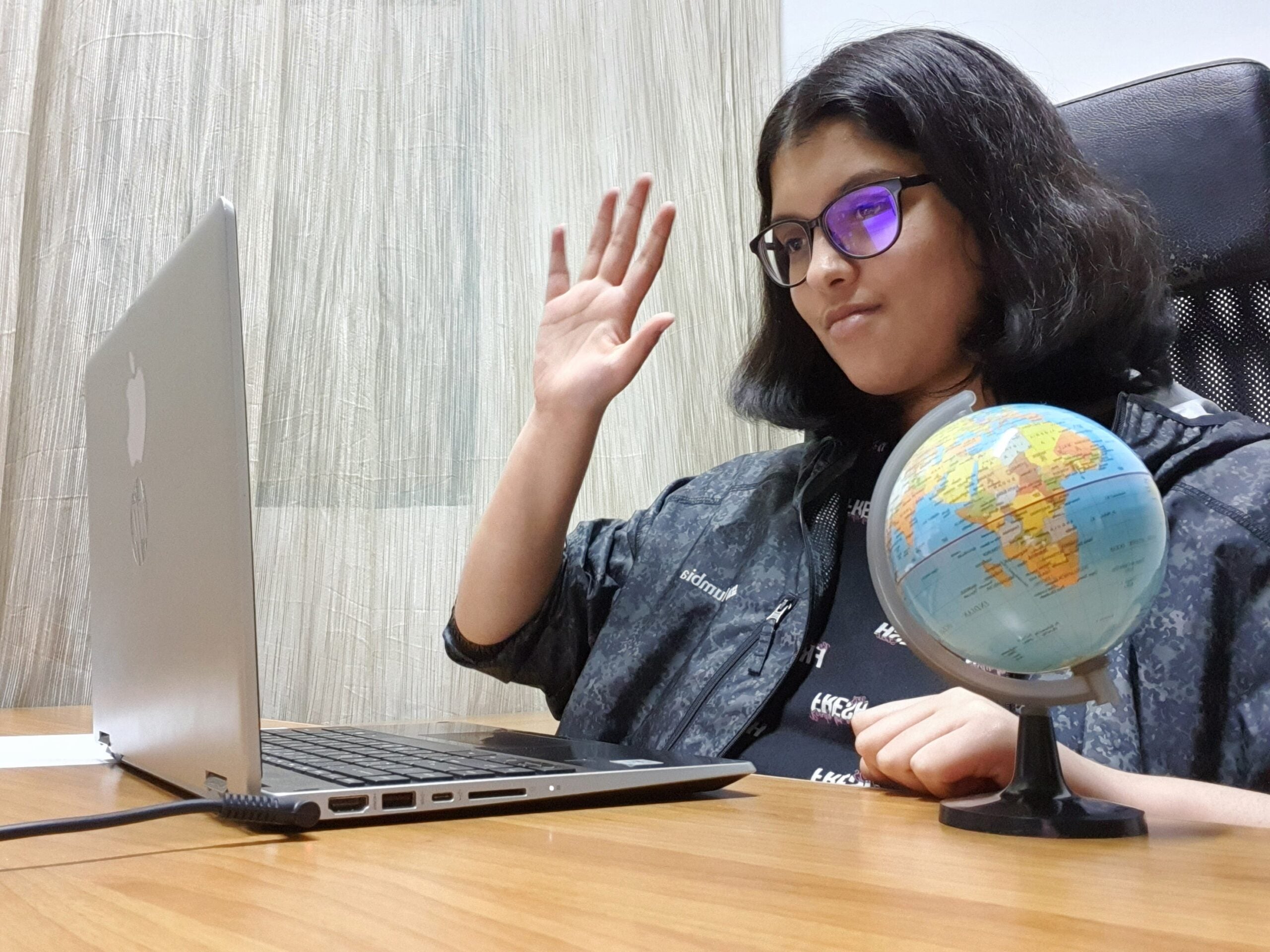 A girl sitting at a desk with a laptop open, waving to the camera. On the desk, there is a small globe.