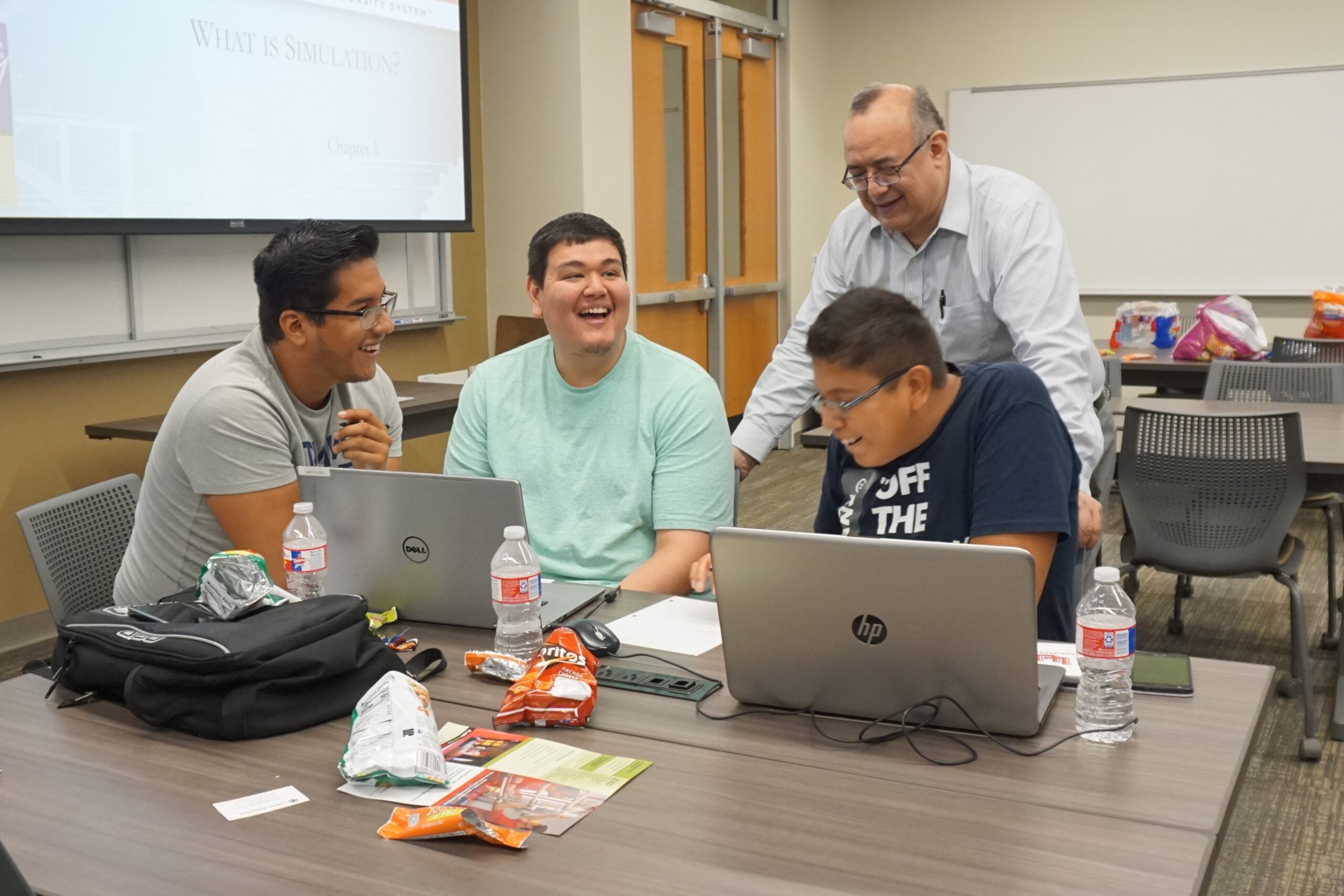 Three students working on laptops at a desk with a teacher behind them