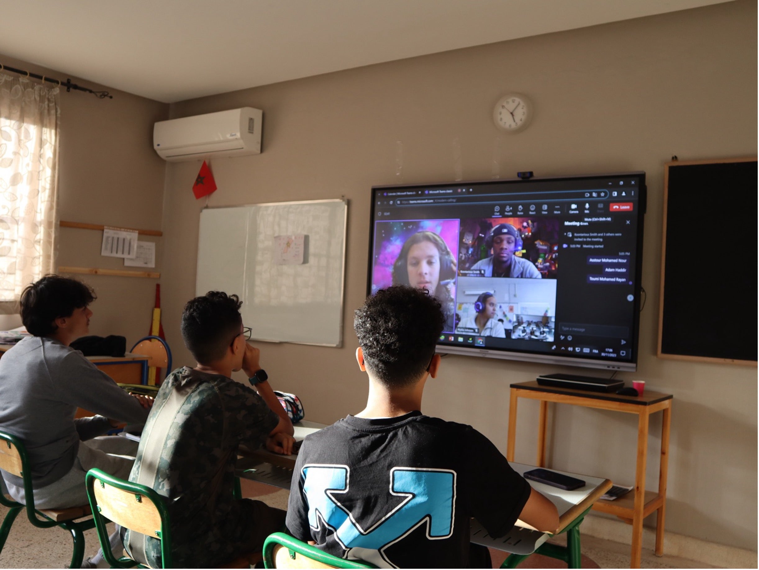 Three students sitting at desks in a classroom, facing a large screen displaying a Zoom meeting with other students.